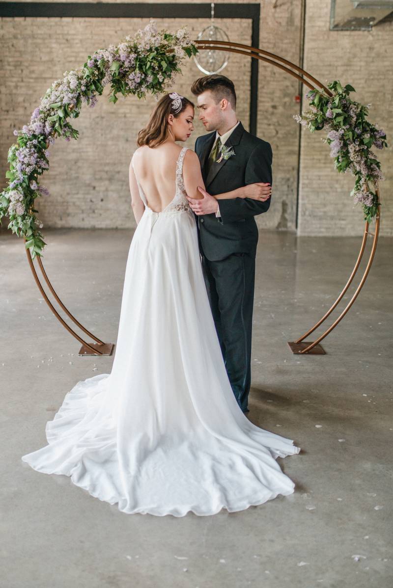 Man and woman embrace looking into eyes in front of copper wedding arch with lilac accents