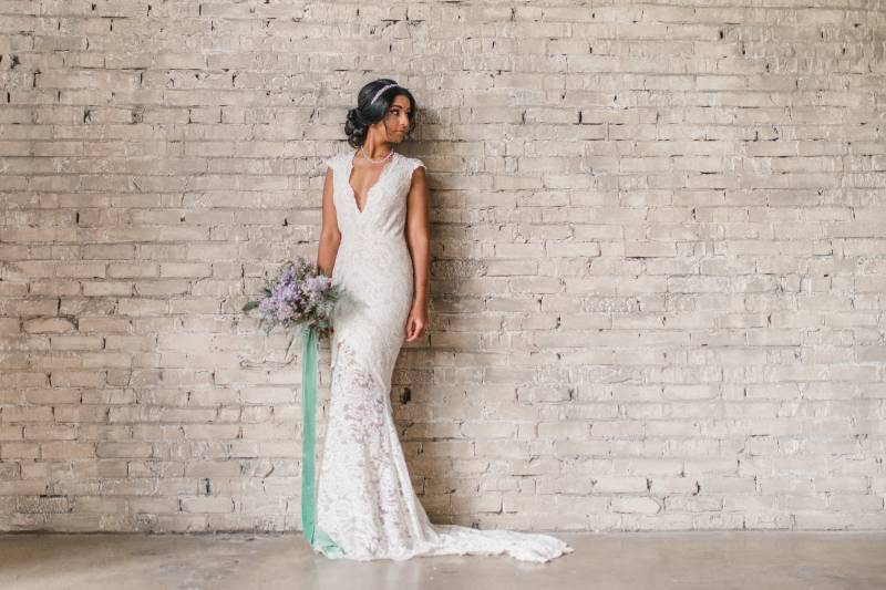 Woman in white lace dress stands looking to side holding lilac bouquet with aqua ribbon 