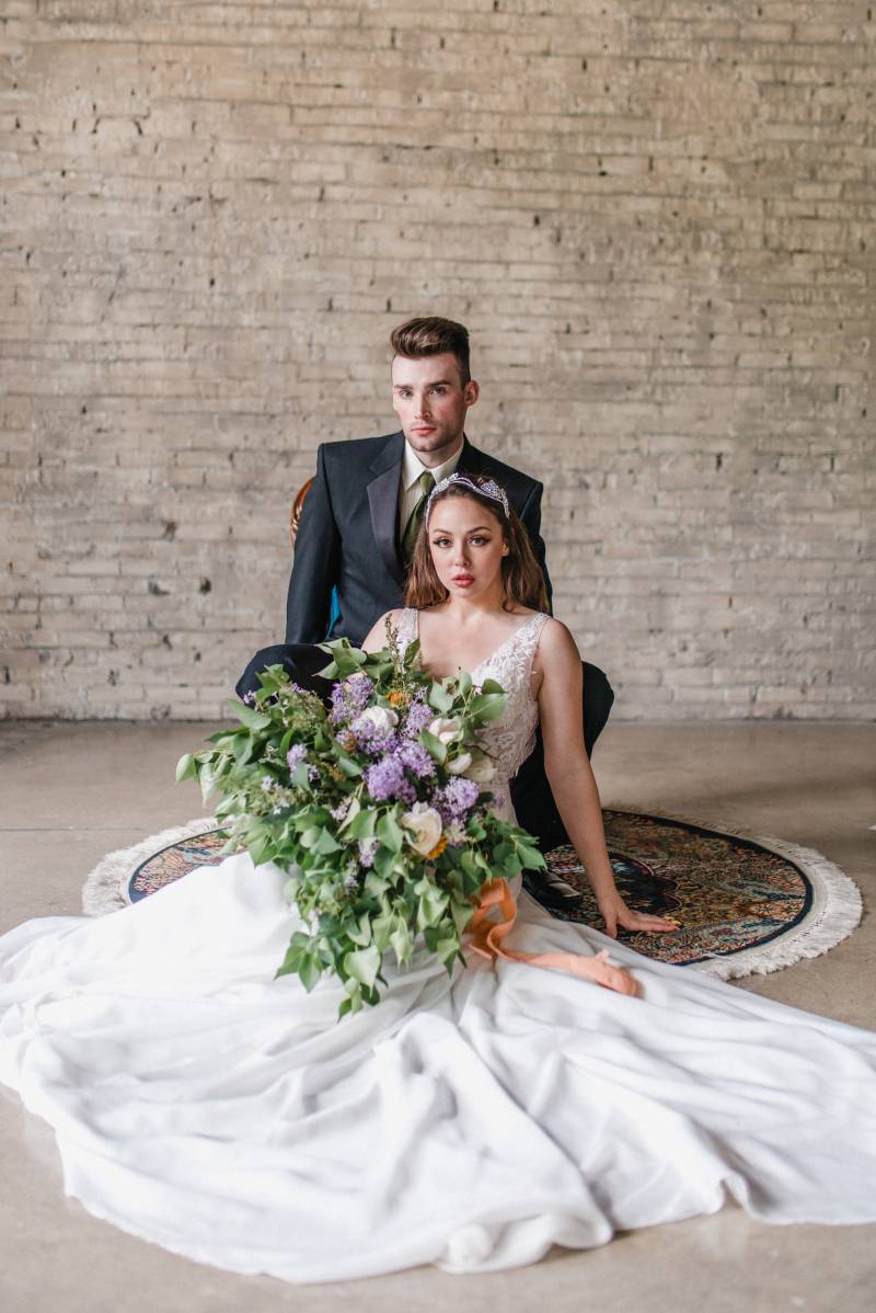 Man sits in black suit jacket looking over shoulder of woman in white lace dress holding lilac bouquet 