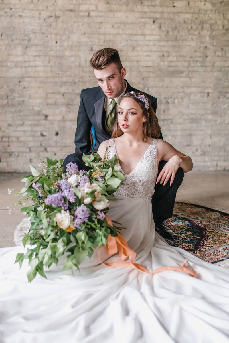 Man sits in black suit jacket looking over shoulder of woman in white lace dress holding lilac bouquet 