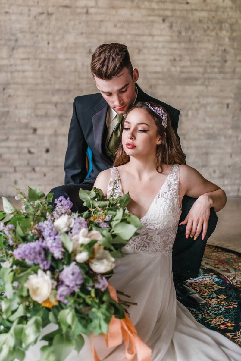 Man sits in black suit jacket looking over shoulder of woman in white lace dress holding lilac bouquet 