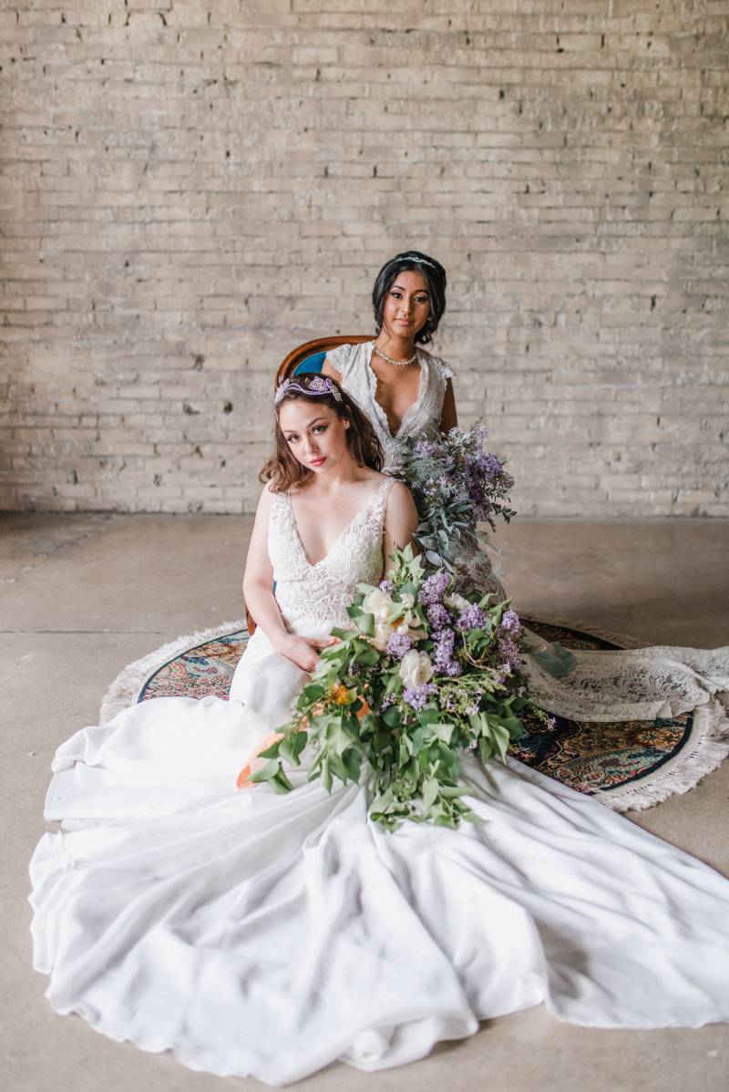 Two woman sit in white lace dresses holding lilac bouquets and blue ribbons 