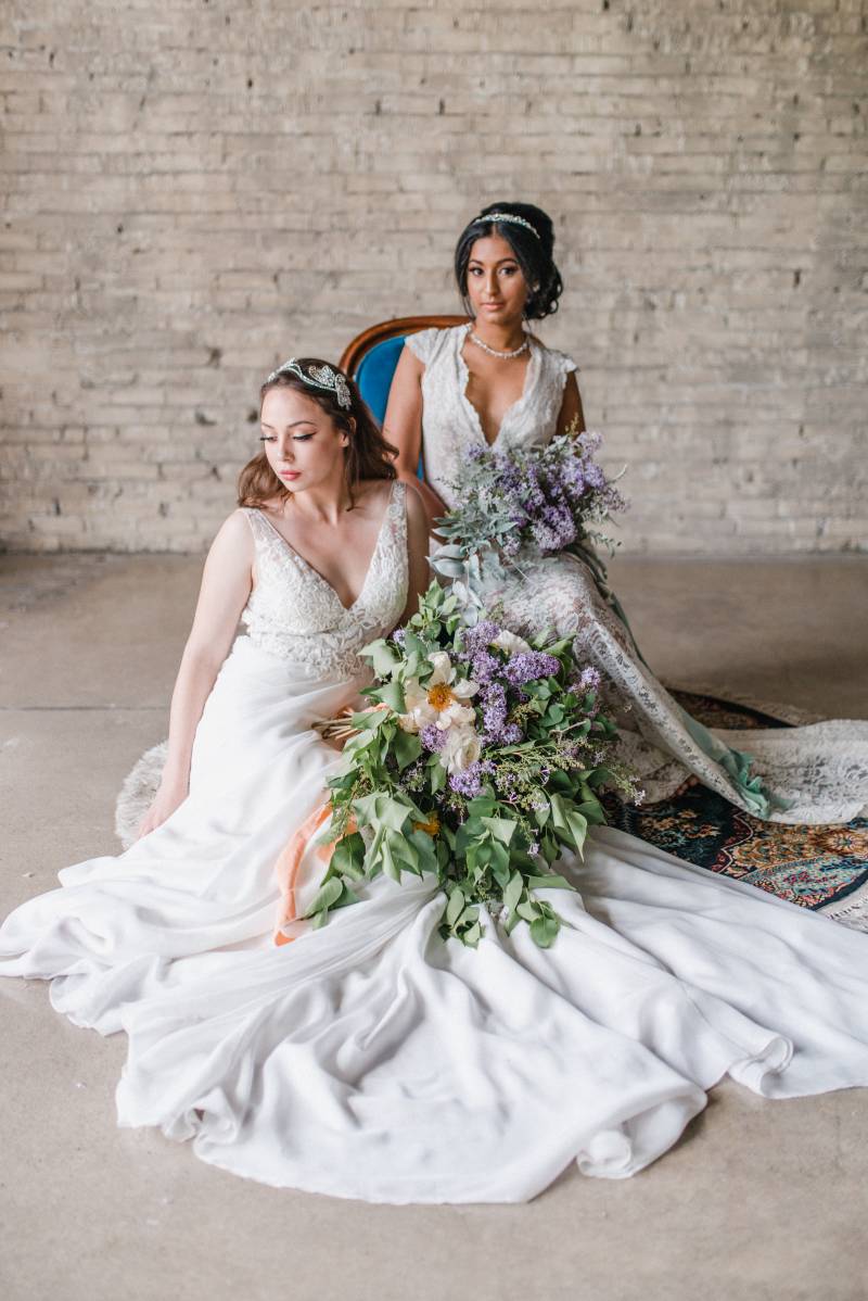 Two woman sit in white lace dresses holding lilac bouquets and blue ribbons 