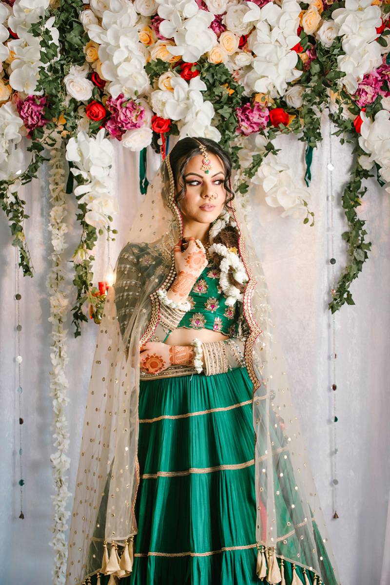 Woman in green dress and clear veil stands in front of flower wall with henna tattoos on wrist and arms