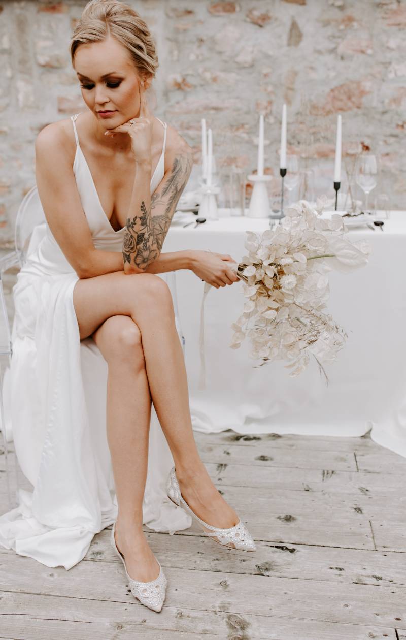 Woman sitting crossed legged in white slit skirt and silver heels holding clear white bouquet 