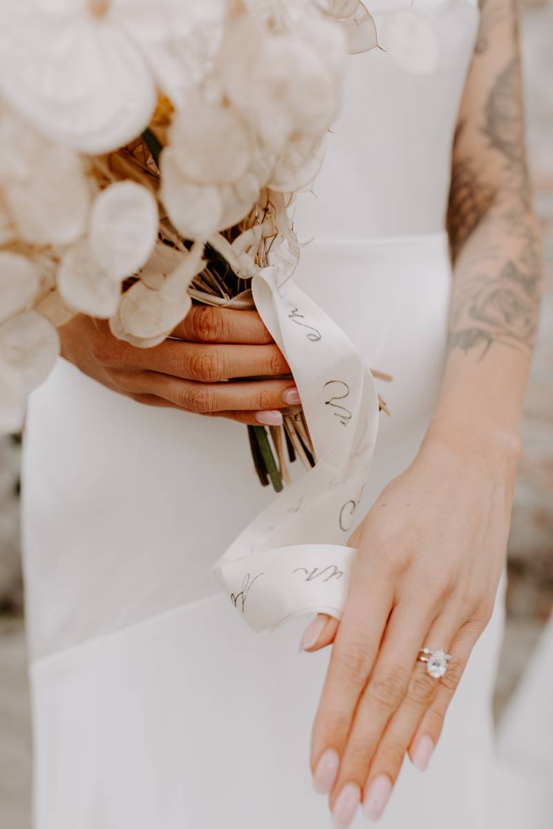 torso of woman in white dress holding out engagement ring and holding clear white bouquet with white ribbon