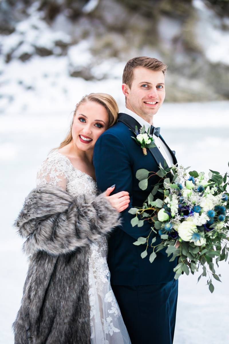 Bride holds groom from behind wearing fur overcoat while groom holds bouquet 