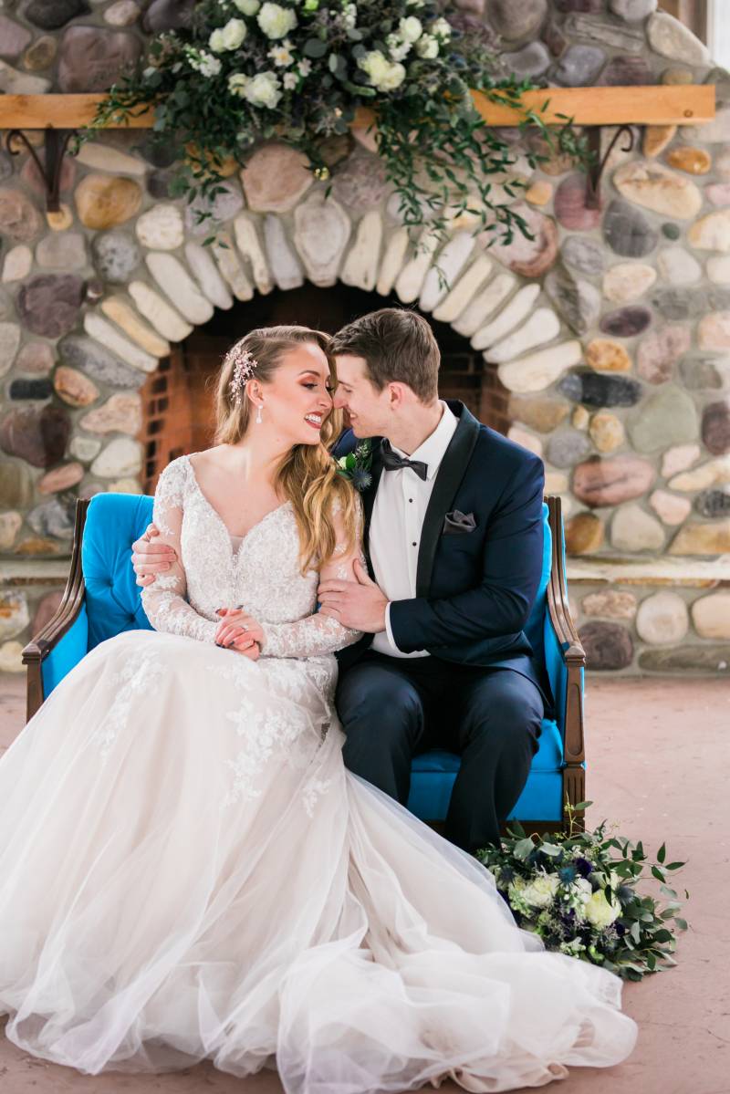 Bride and groom sit on blue sofa embracing smiling in front of store fireplace 
