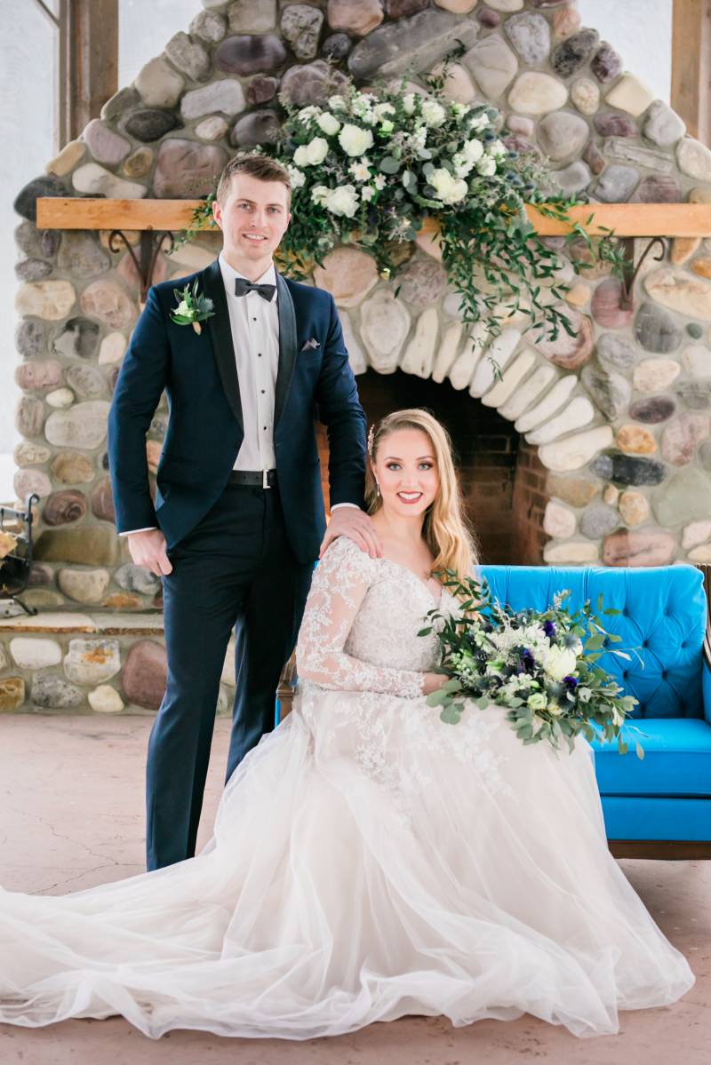 Groom stands with hand on shoulder of Bride in white lace sleeved dress holding white blue and purple bouquet 