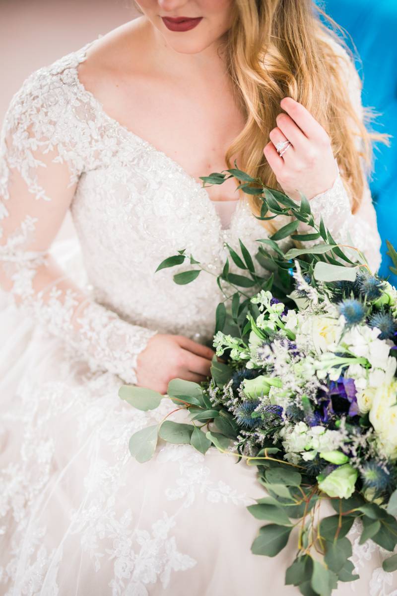 Bride sits in white lace sleeved dress holding white blue and purple bouquet 