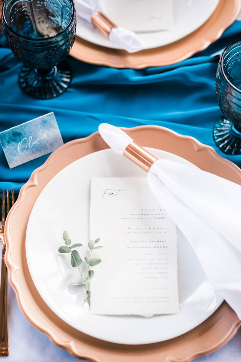 Place setting flat lay with menu on bronze plate and bronze cutlery on blue table cloth