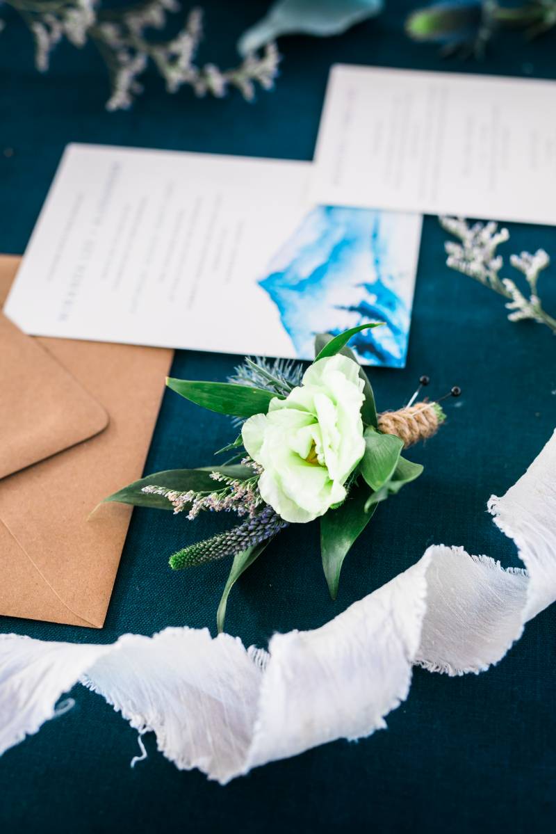 Lime green boutonniere laying on blue table with blue wedding invitation behind  