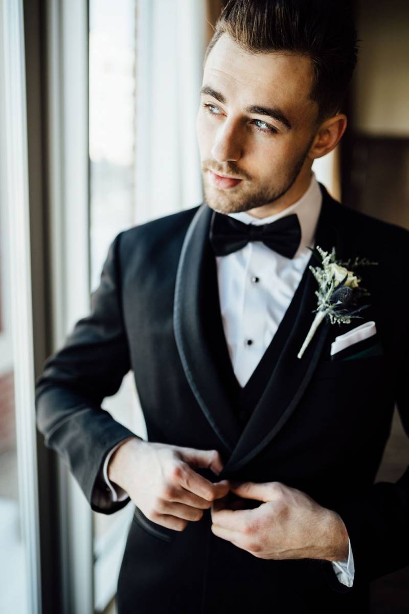 Man buttons up black suit jacket with boutonniere while looking towards window 