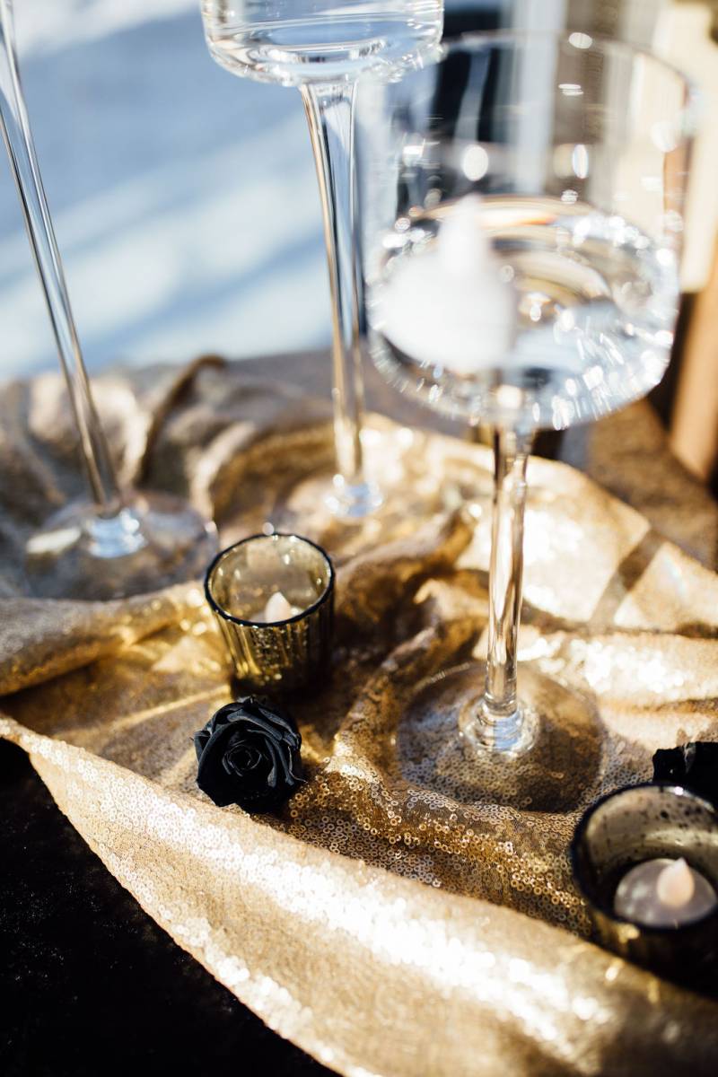 Tall wine glasses on top of sparkling gold table runner with black roses 