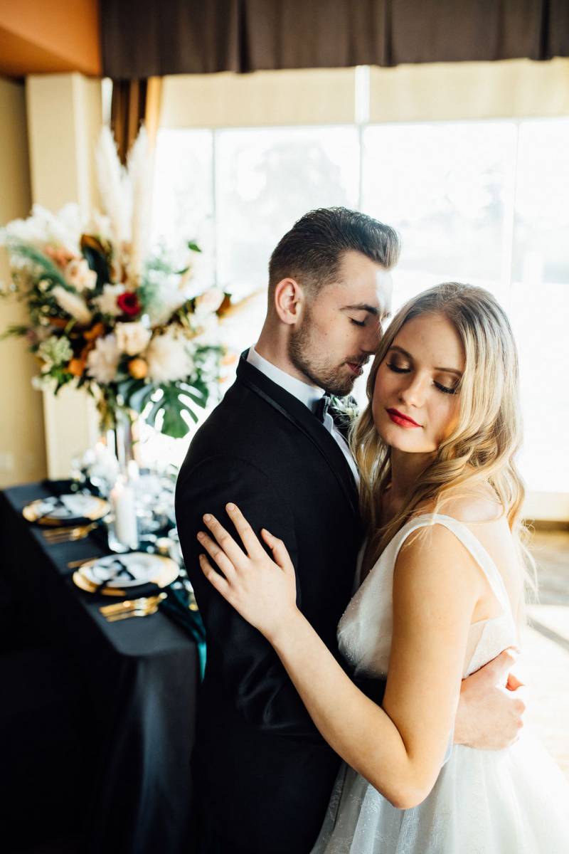 Bride and groom embrace in front of black table with large floral centerpiece 