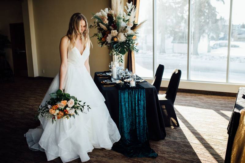 Bride in large white dress standing beside table with large floral centerpiece holding orange and pink bouquet 