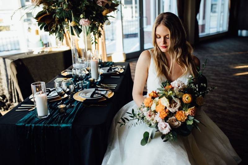 Bride in white dress sitting holding orange and blush bouquet beside table 
