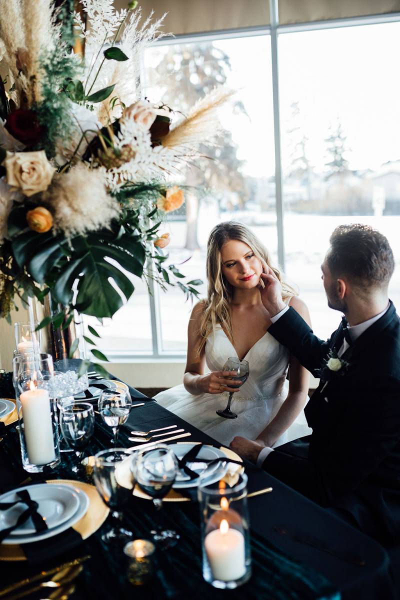 Groom touches Woman's face sitting at black table with large floral centerpiece 
