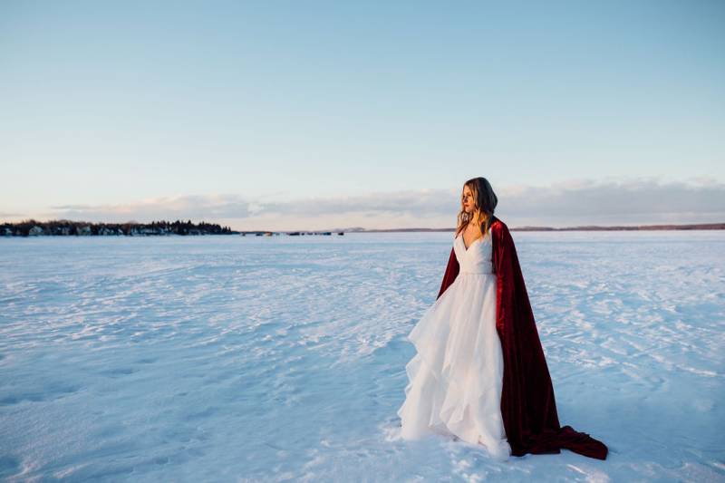 Woman in large white dress and red cape standing in large open snowy field 