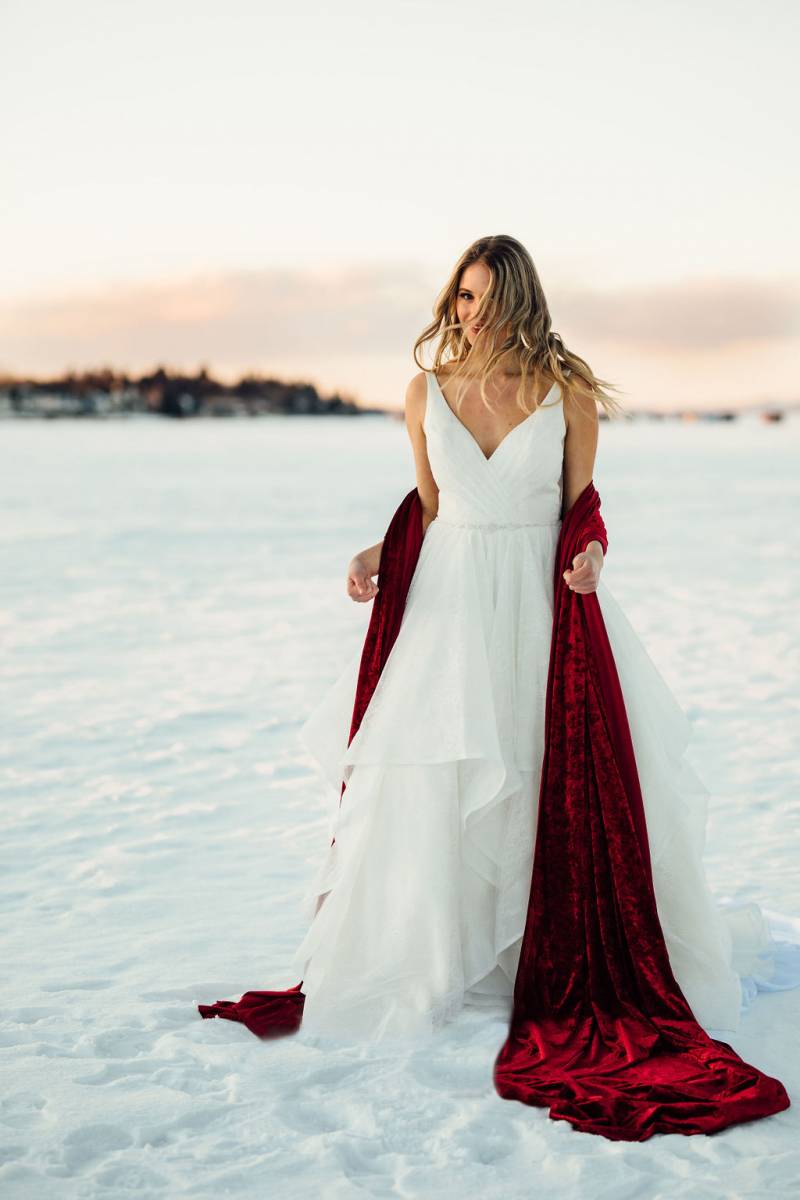 Woman in large white dress wrapped in velvet red fabric standing in large open snowy field 