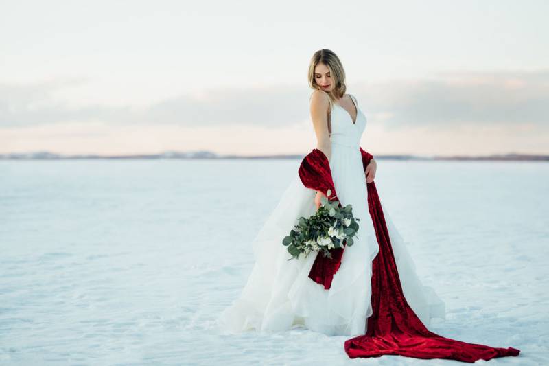 Woman in white dress holding white floral bouquet wrapped in red velvet fabric in snowy field 