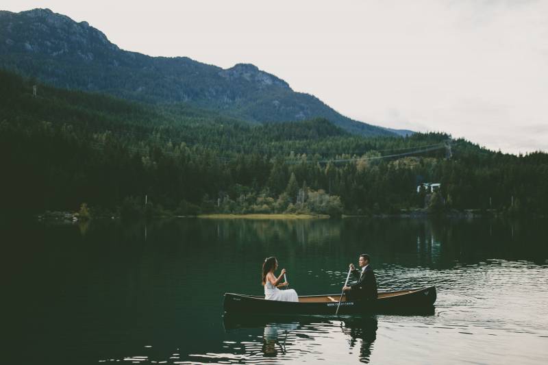 Bride and groom sit facing paddling out on green lake in canoe with mountain background 