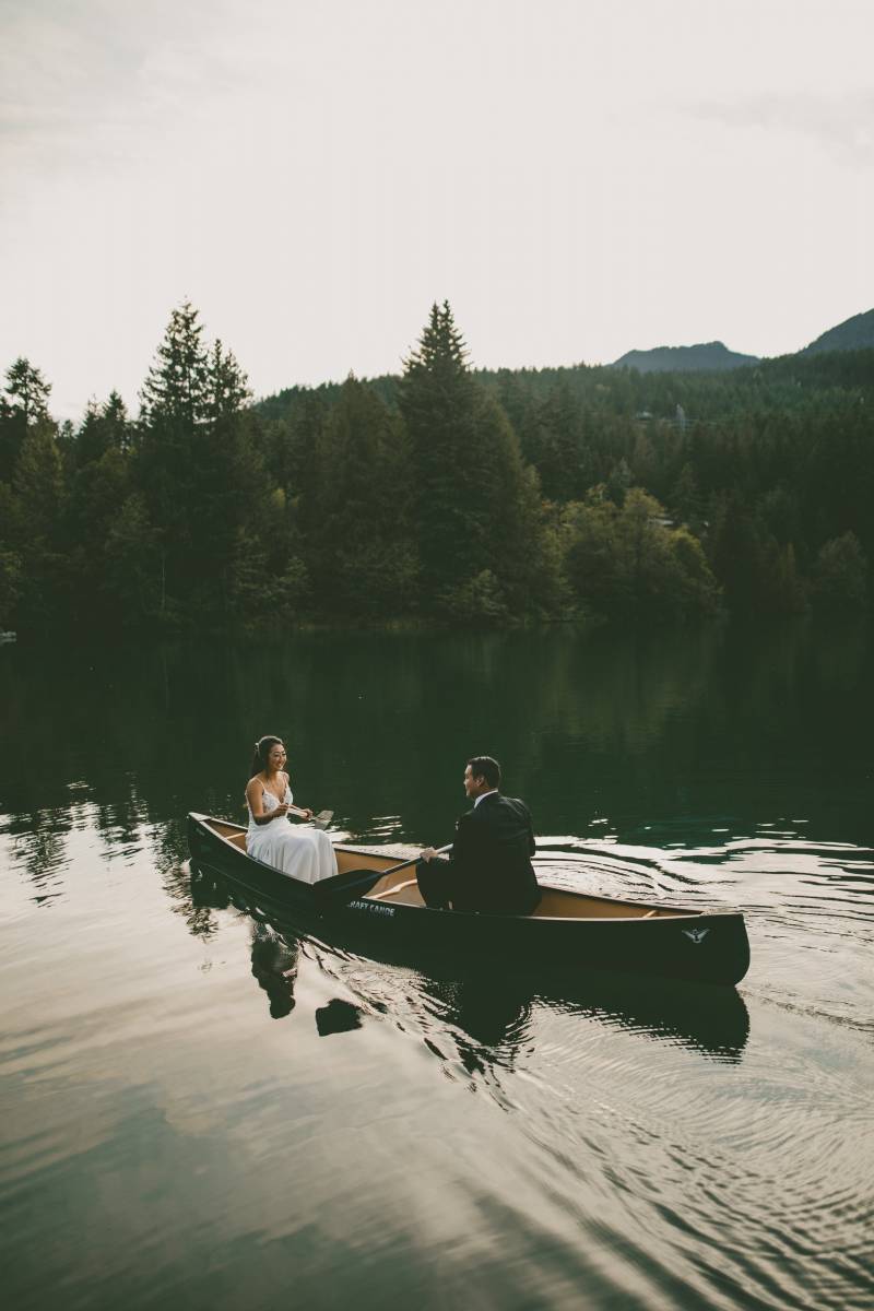 Bride and groom sit facing paddling out on green lake in canoe with mountain background 