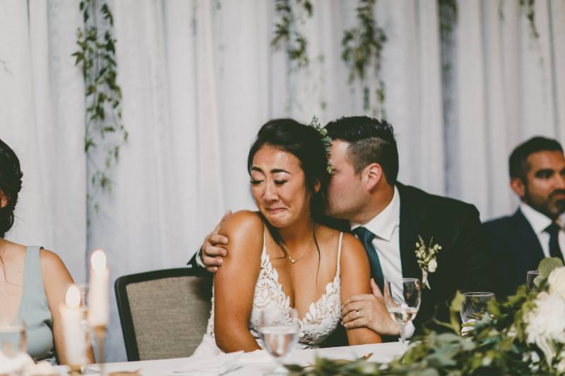 Groom kisses back of smiling bride sitting holding shoulder 