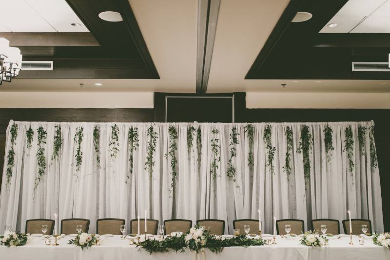 Head table with white floral centerpiece and white handing fabric with handing greenery 