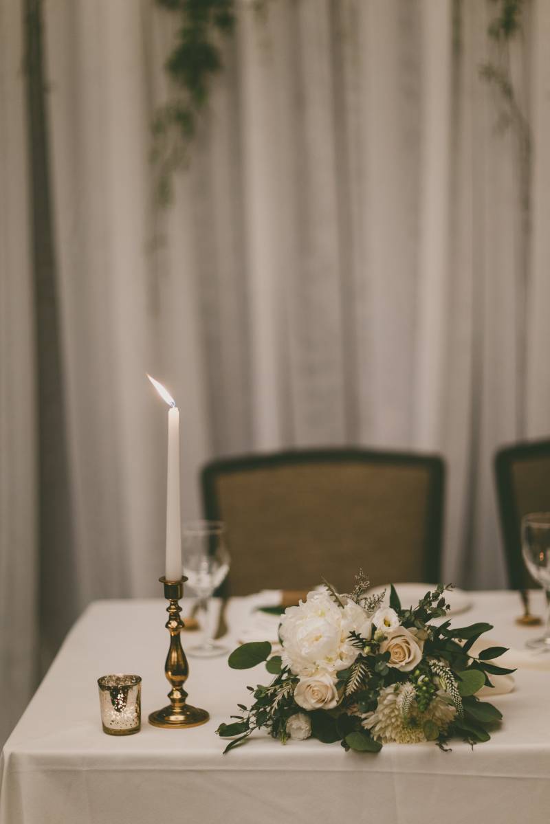 Lit candlestick at edge of white table beside white floral centerpiece 