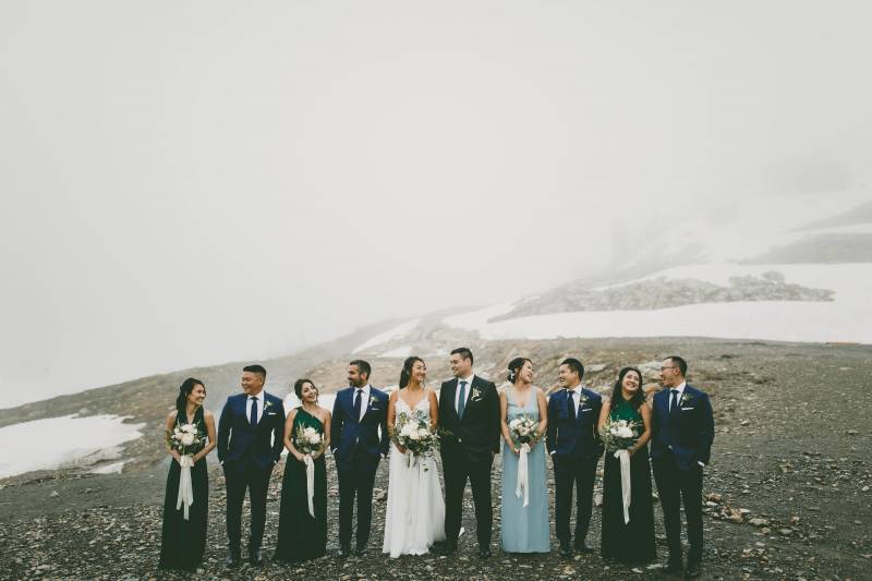 Bridesmaids and groomsmen stand beside groom and bride atop foggy gravel mountain 