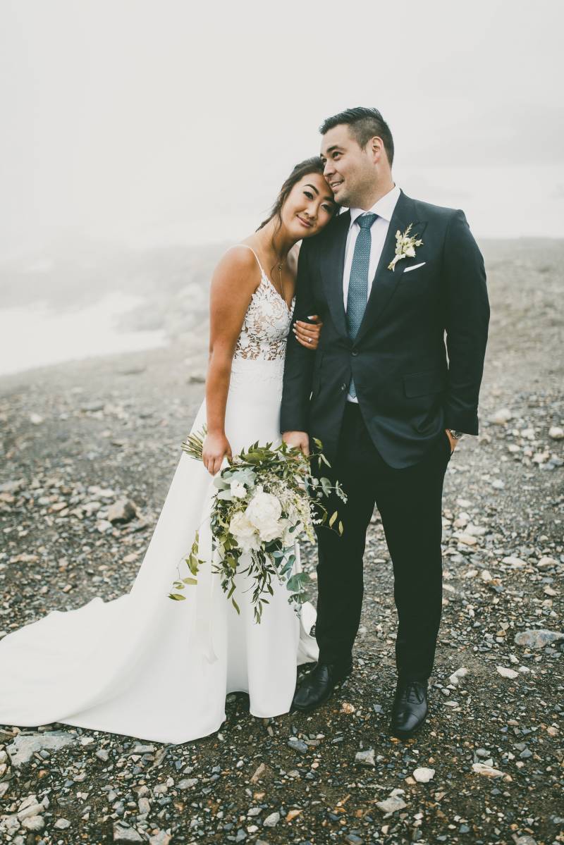Bride and groom stand leaning on shoulder holding bouquet on gravel while foggy 