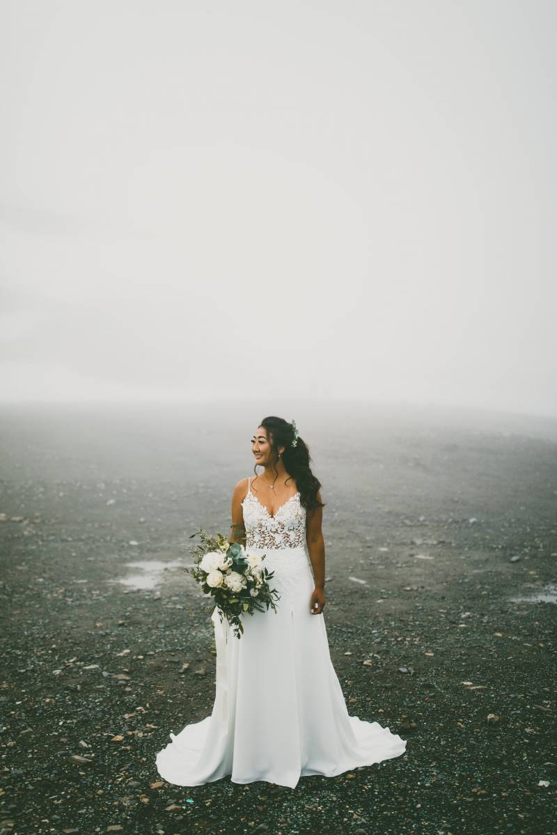 Bride in white dress holding white bouquet stands on foggy gravel 