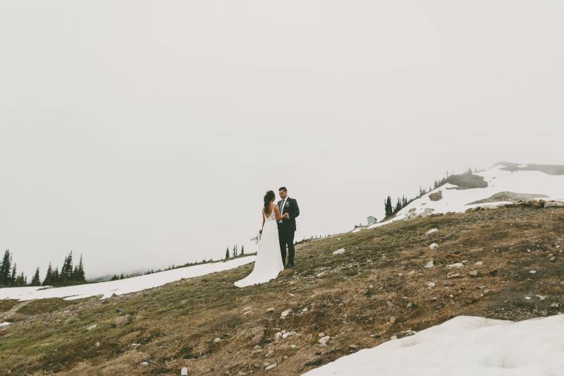 Bride and groom stand holding hands on snowy and grassy ledge 