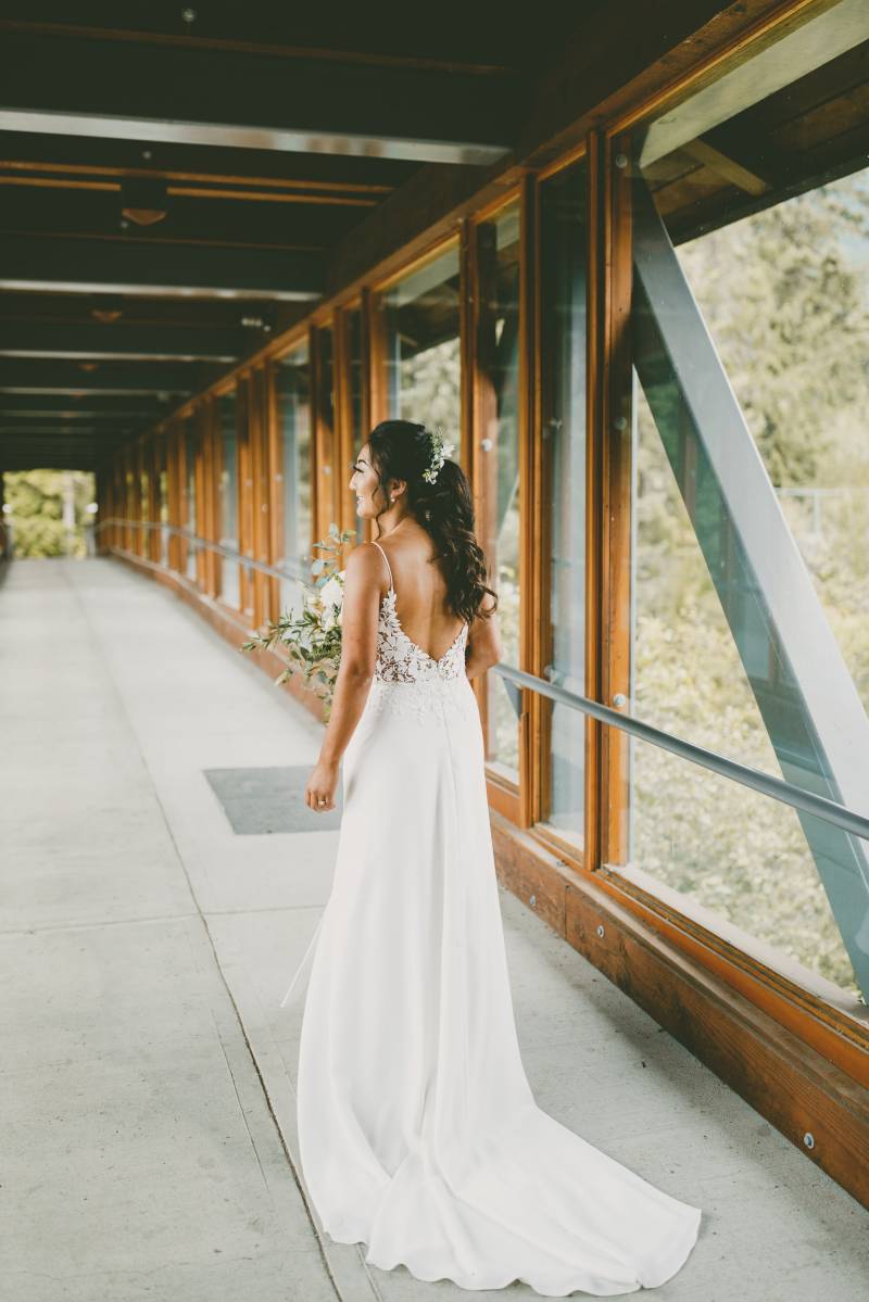 Bride in long white open back dress walks down windowed hallway 