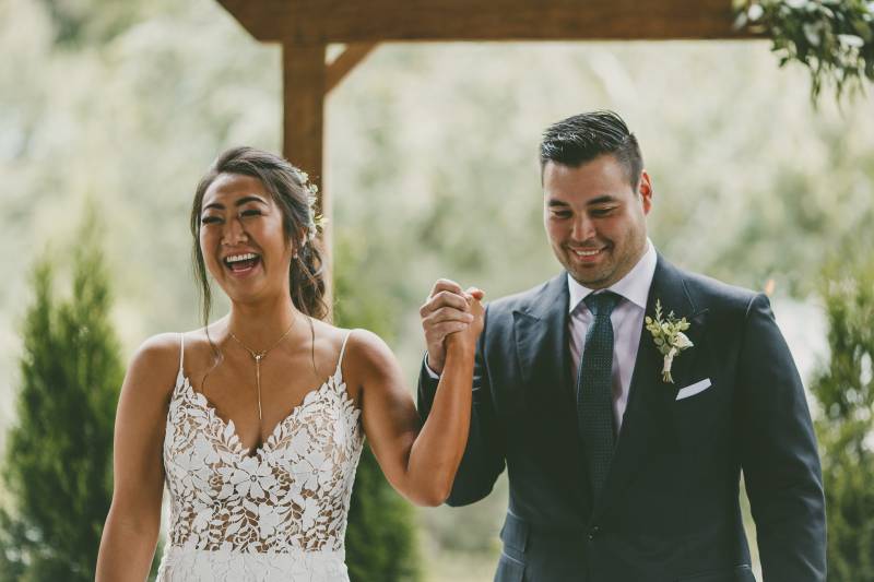 Bride and groom stand holding hands raised while laughing 
