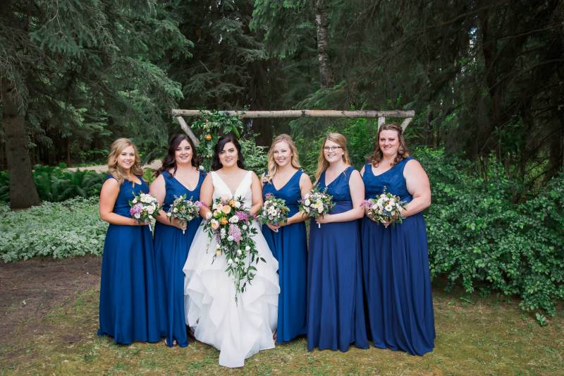 Bride and bridesmaids stand holding bouquets in front of wooden wedding arch 