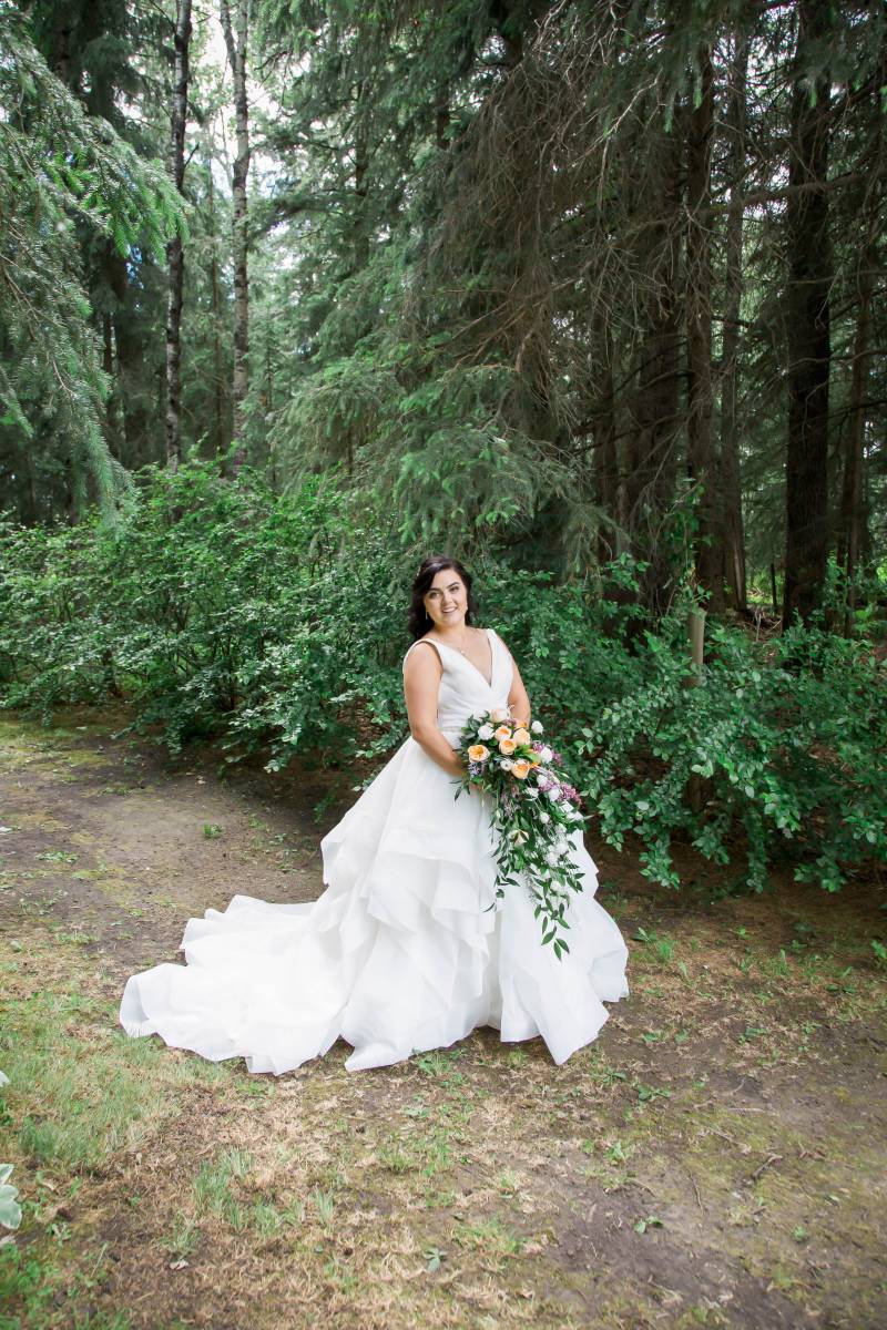 Bride holding yellow purple and white bouquet stand in front of dark green forest 