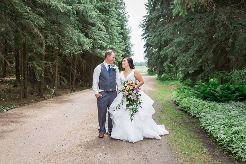 Bride and groom smiling holding hands on dirt pathway between forest