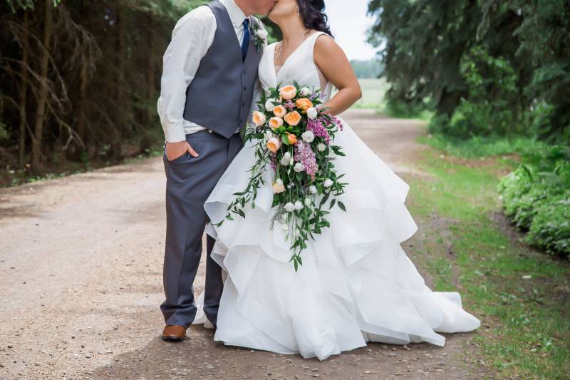 Groom and bride kiss holding yellow white and purple bouquet on dirt pathway