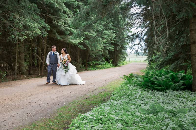Bride and groom walk holding hands down dirt road between dark forest