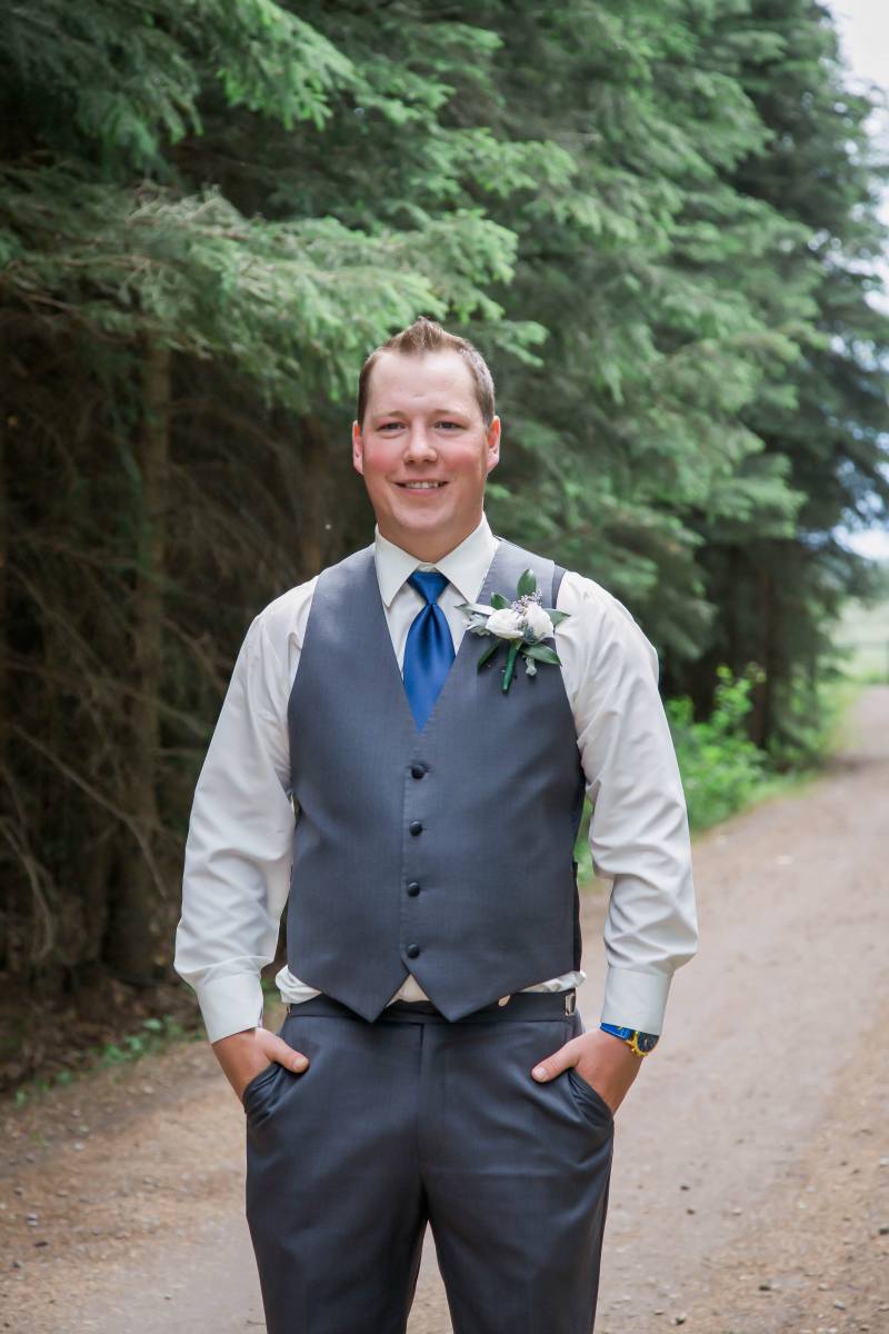Groom stand on dirt road smiling with hands in pockets
