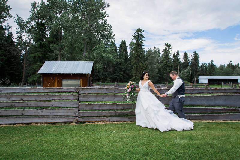 Bride and groom walking holding hands beside wooden fence on grass 
