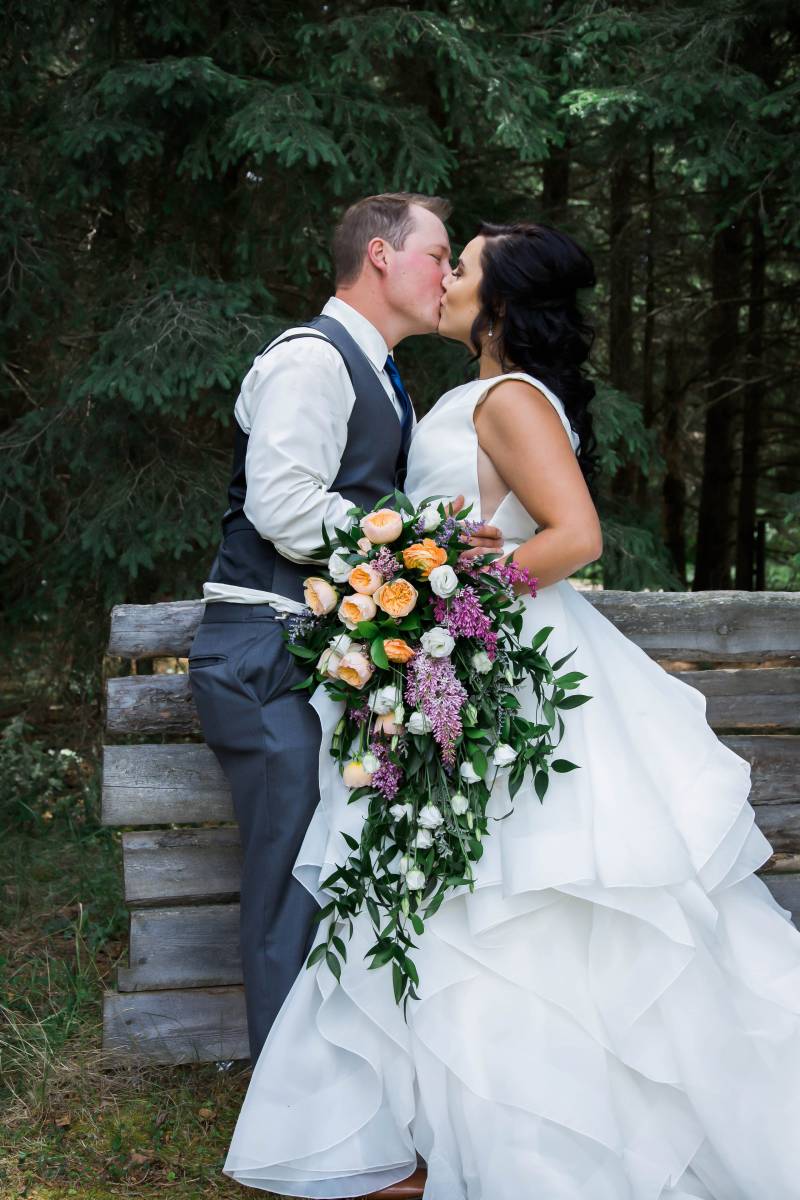Bride kisses groom holding yellow purple and white bouquet in front of dark green forest