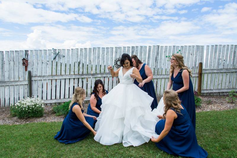 Bridesmaids hold brides white dress above ground in front of white picket fence 