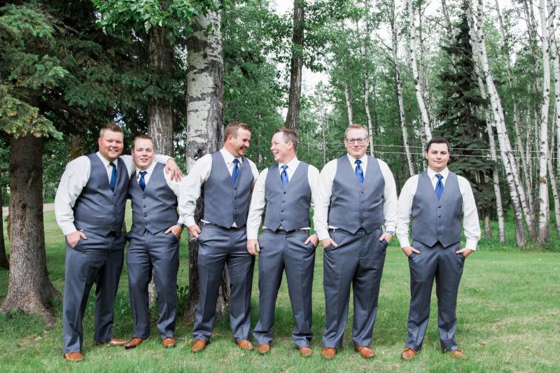 Groomsmen and groom stand together hands in pockets smiling 