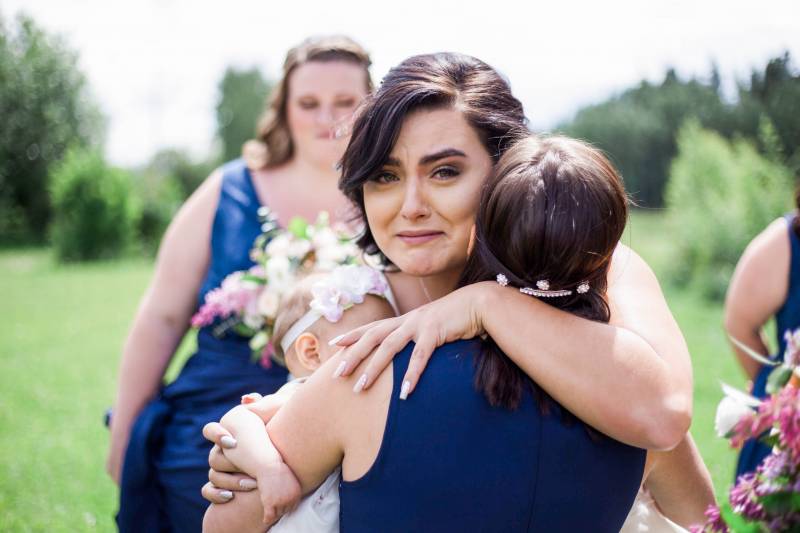 Bride smiling while embracing bridesmaid and holding child in white dress