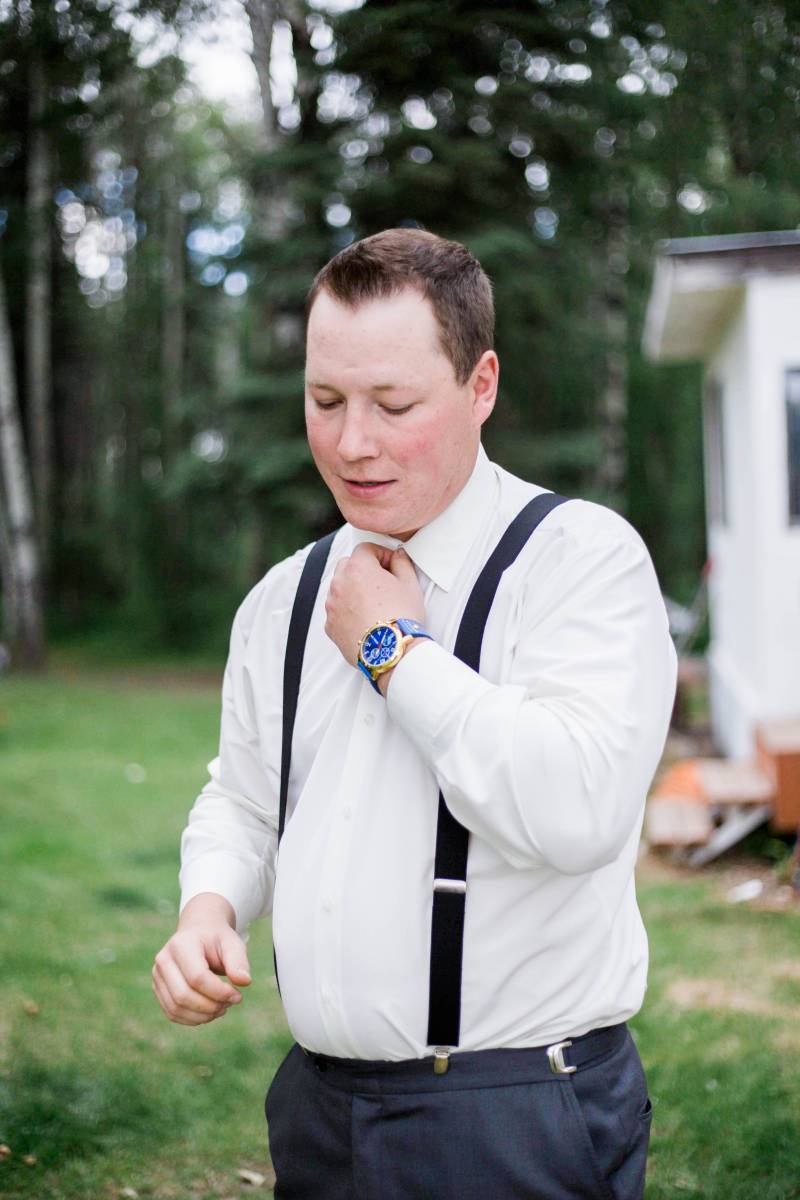 Groom in white shirt and black suspenders adjusts neck tie