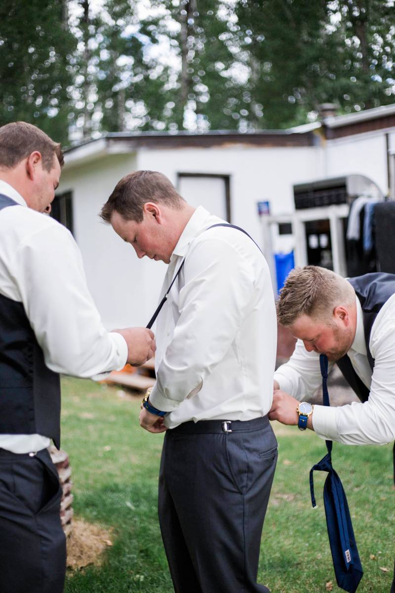 Groom and groomsmen help adjust suit and pants outside 