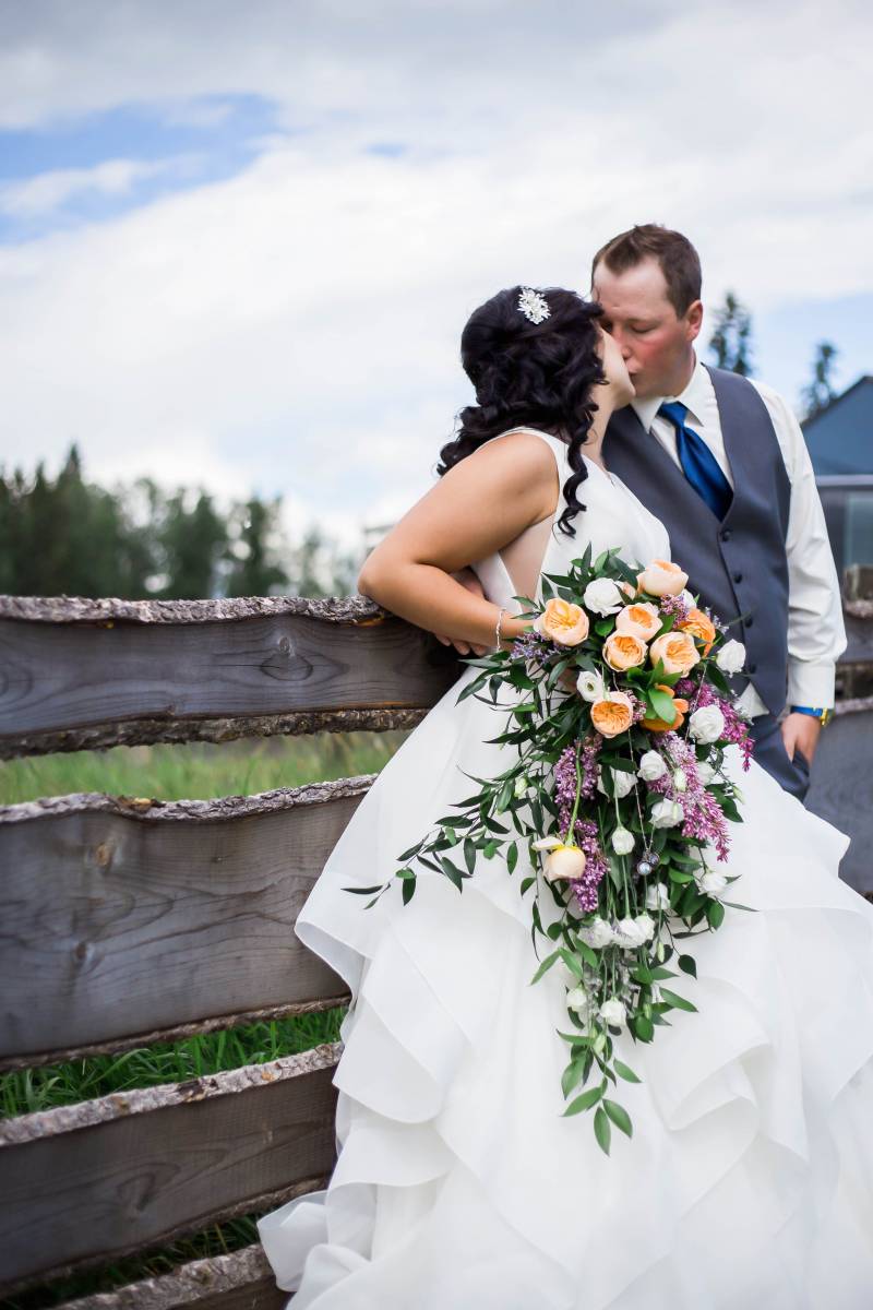 Bride holding yellow purple and white bouquet kissing groom leaning on old wooden fence